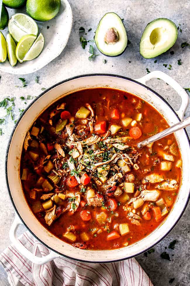 top view of a big white pot of Caldo de Pollo (Mexican Chicken Soup) with limes and avocados ready to garnish