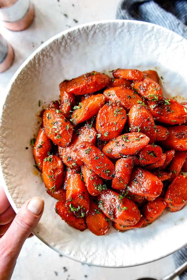a hand holding a white bowl of brown sugar glazed carrots