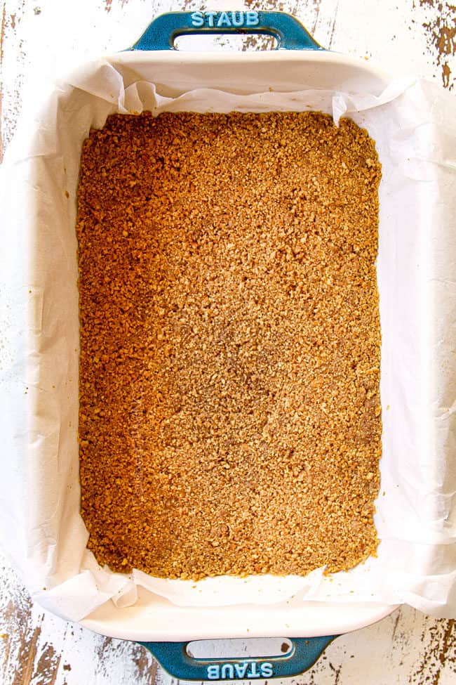showing how to make pumpkin cheesecake bars by pressing crust crumbs into a 9x13 baking dish