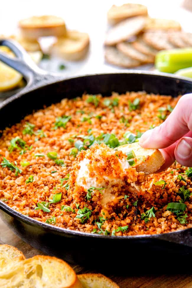 a hand dipping into baked crab dip in a black skillet with a baguette slice
