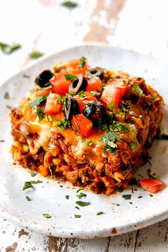 showing how to top Mexican Lasagna recipe by adding sour cream, olives, tomatoes and cilantro to a piece of lasagna on a plate