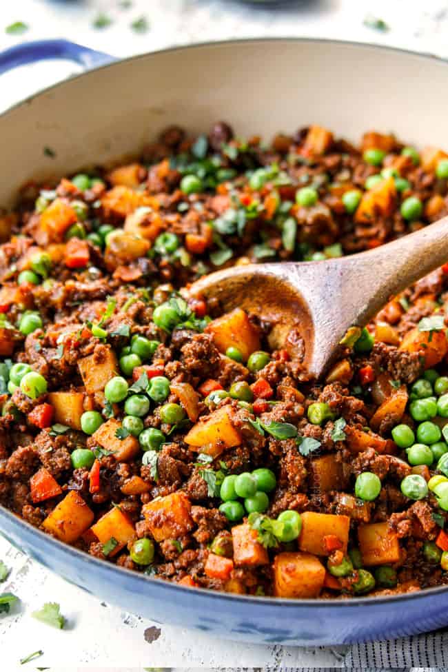 showing how to make Mexican Picadillo recipe by stirring ground beef, potatoes, carrots and peas together in a blue pot with a wooden spoon
