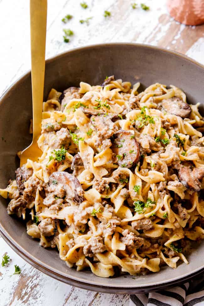showing how to serve beef stroganoff with ground beef by placing in a bowl with egg noodles, garnished with parsley with a gold fork