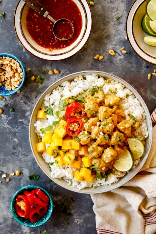 showing what to serve with Thai peanut chicken by showing chicken in a bowl over rice with garnish bowls of chili sauce, peanuts, limes