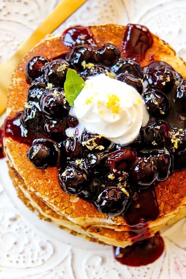 top view showing how to serve lemon ricotta pancakes by stacking pancakes on a white plate with blueberry sauce and a dollop of whip cream