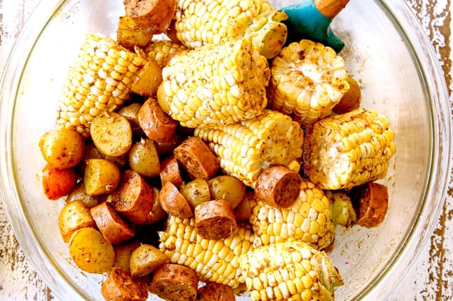 showing how to make shrimp boil by combining corn, potatoes, and sausage in a glass bowl with olive oil, Old Bay Seasoning and ground mustard and tossing to coat.