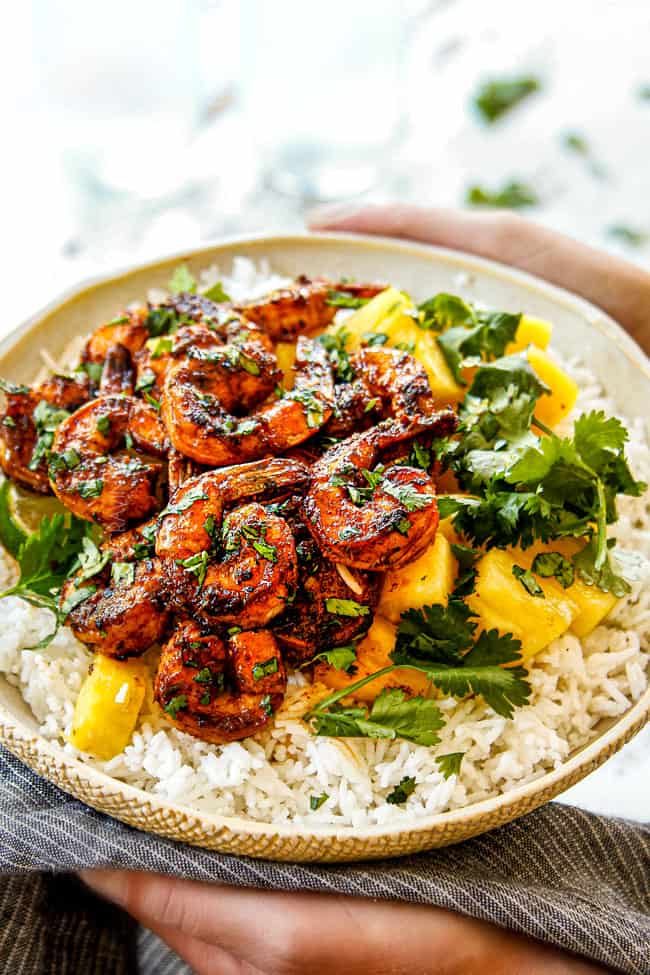 two hands holding a bowl of jerk shrimp with rice and pineapple