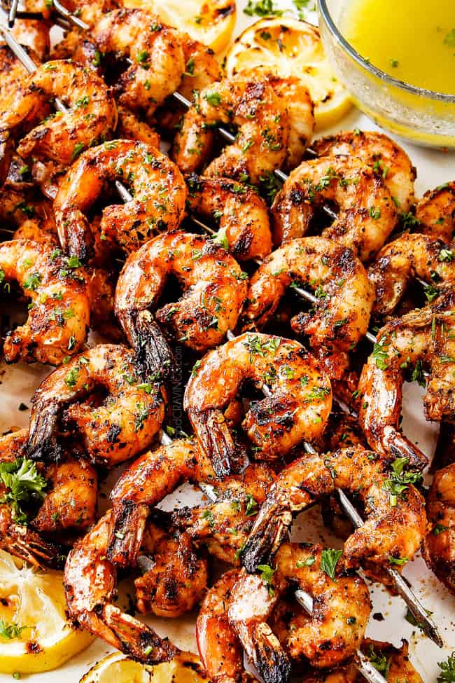 a stack of lightly charred grilled shrimp skewers on a white plate garnished by parsley