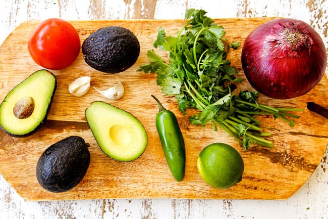 showing how to make best guacamole recipe by laying out ingredients on a wood cutting board: avocados, tomatoes, garlic, jalapenos, cilantro, lime and red onion