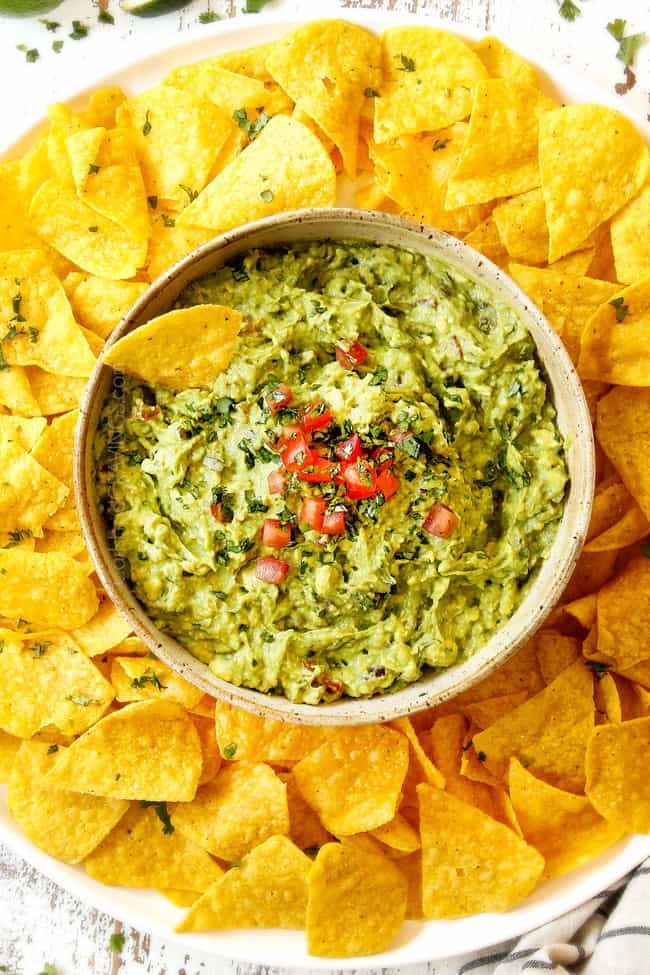 showing how to garnish guacamole recipe by topping with fresh tomatoes in a tan bowl surrounded by tortilla chips
