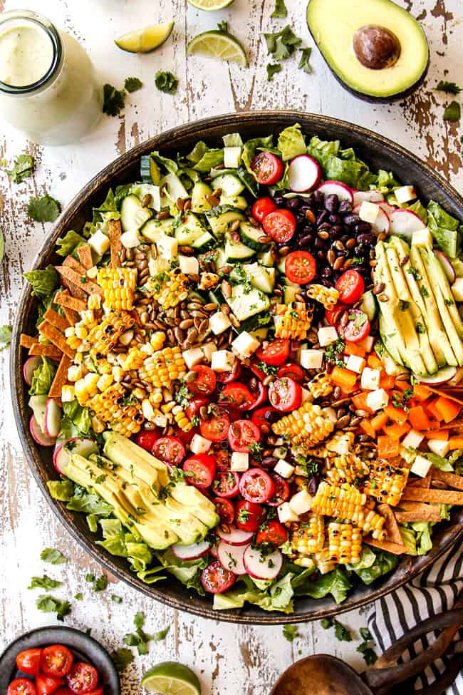 top view showing how to make Mexican Salad recipe by adding tomatoes, corn, black beans, cucumbers and avocados to a wooden bowl and garnishing with cheese, pepitas and tortilla strips