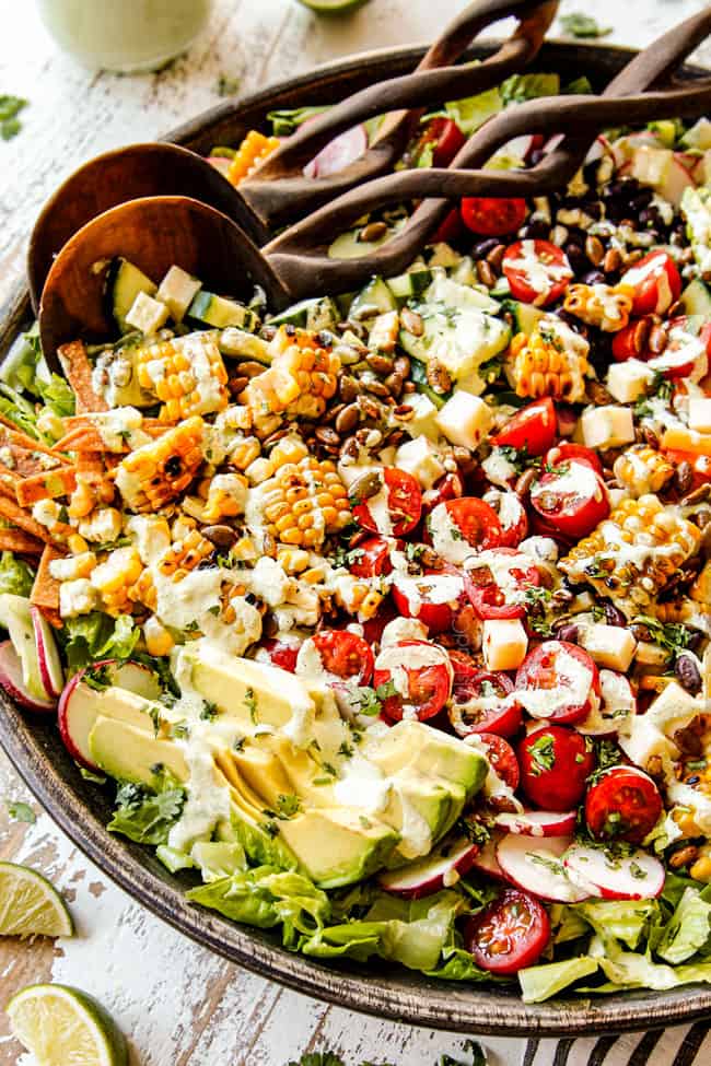 up close of Mexican Salad in a wooden bowl with wooden serving spoons showing the ingredients: avocados, corn, tomatoes, radishes and avocados