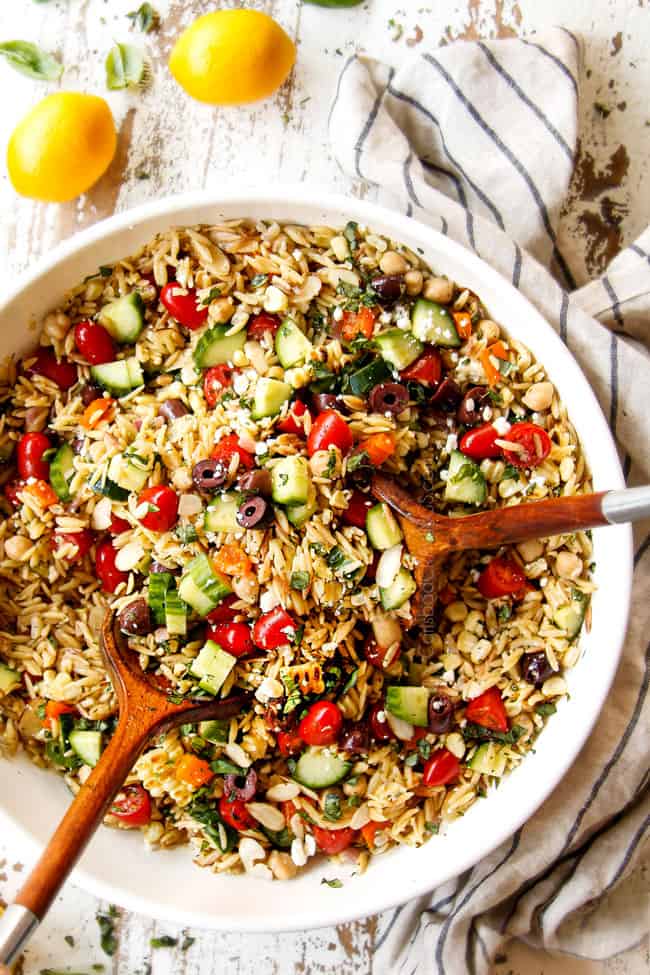 showing how to make Greek orzo pasta salad by adding tossing orzo, cucumbers, bell peppers, tomatoes, olives, chickpeas, basil and feta together with dressing in a white bowl