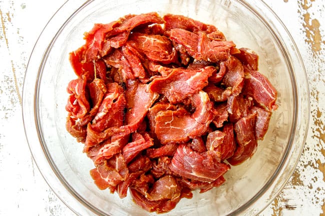 showing how to make Pepper Steak by marinating slices of flank steak in a glass bowl with soy sauce, sesame oil, rice wine, Chili sauce and cornstarch