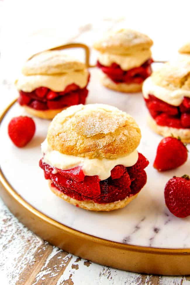 showing how to serve strawberry shortcake by layering biscuits with strawberries, whipped cream and topping with half a biscuit and placing on a white marble platter