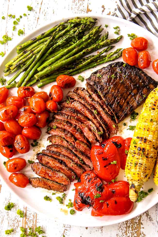 top view of flank steak cut in thin slices on a white platter with grilled corn and asparagus and bell peppers showing how to serve