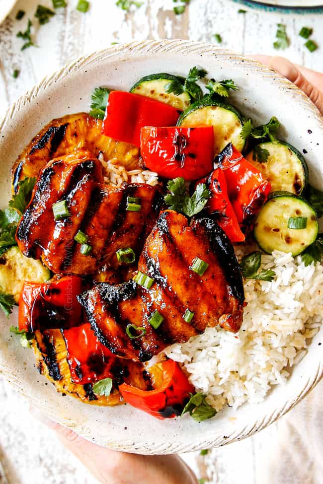 top view of two hands holding a cream plate of Hawaiian chicken with white rice, grilled bell peppers and zucchini