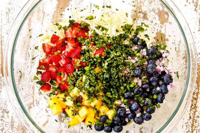 showing how to make fish tacos by making slaw in a glass bowl with cabbage, cilantro, strawberries and pineapple