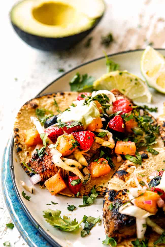 showing how to make mahi mahi fish tacos by adding fish to a corn tortilla and topping with slaw and avocados