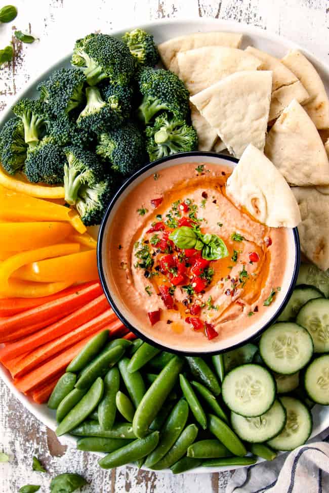 top view of hummus in a black bowl on a platter with carrots, broccoli, cucumbers, snap peas, bell peppers and pita bread