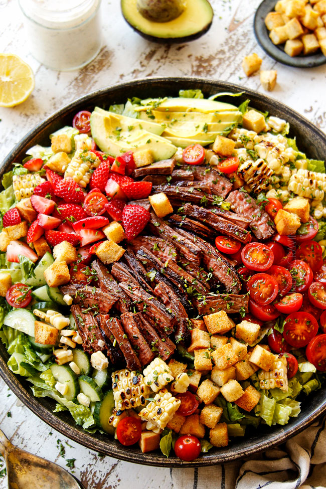 side view of steak salad recipe in a wooden bowl with sliced steak, corn, croutons, tomatoes, avocados and strawberries