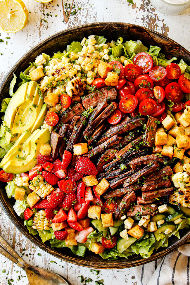 top view showing how to serve steak salad recipe by layering Romaine lettuce with grilled steak, tomatoes, corn, croutons, avocados, cucumbers and strawberries