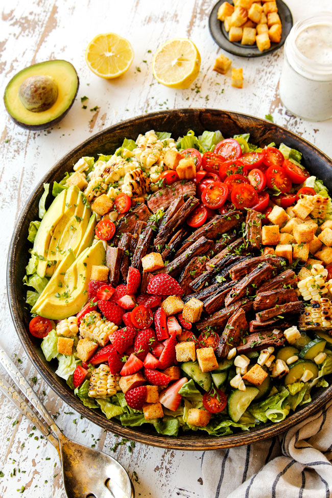 making steak salad by adding lettuce, steak, cucumbers, corn, tomatoes, steak and blue cheese to a wooden bowl