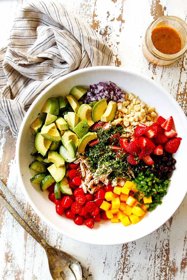 top view showing how to make avocado chicken salad by adding shredded chicken, avocados, corn, black beans, cilantro, red onions, strawberries, mangos and tomatoes to a white bowl