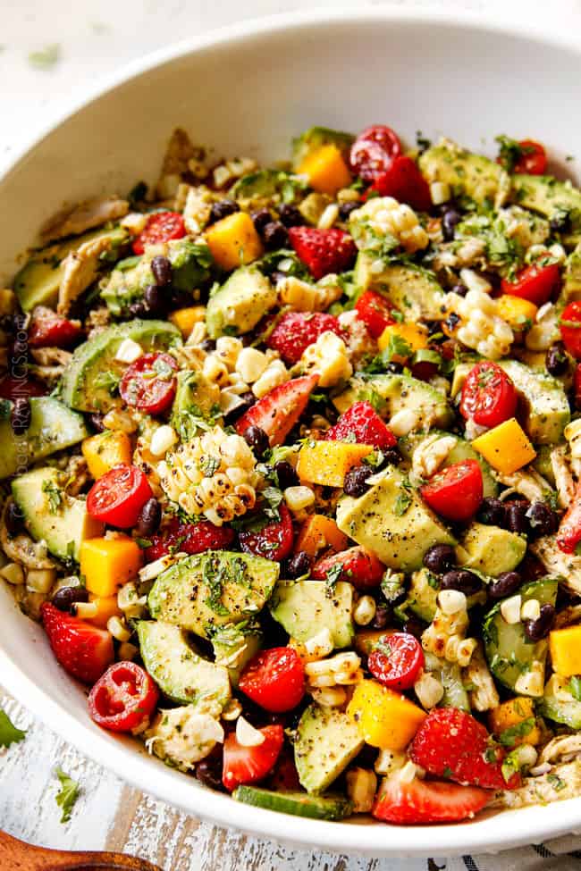 side view showing what's in avocado chicken salad: avocados, corn, black beans, red onions, cilantro, tomatoes, mangos and strawberries in a white bowl