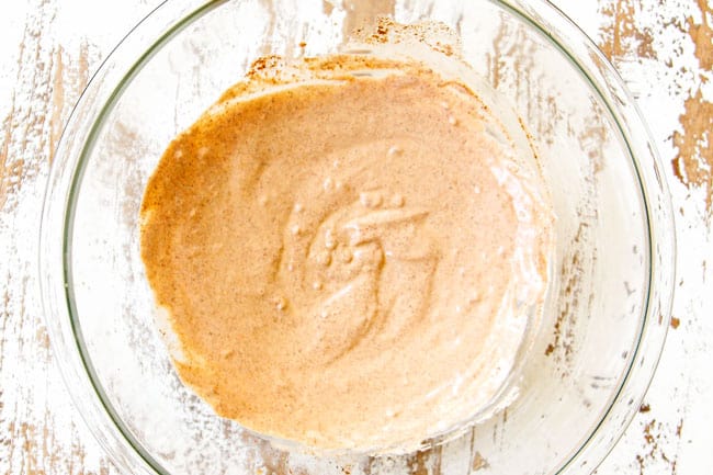 showing how to make Mexican Corn Salad (esquites) by whisking together sour cream, mayonnaise, cotija cheese, chili powder, salt, pepper and ground cumin in a glass bowl to make the dressing