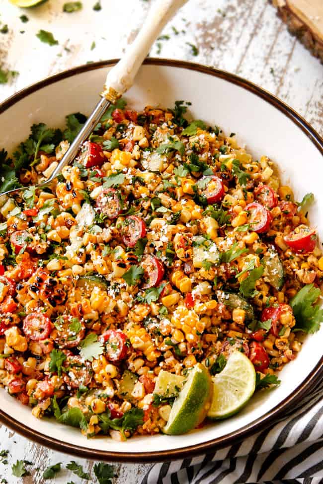 side view of Mexican Corn Salad (esquites) in a white bowl garnished with cotija cheese, cilantro and lime wedges