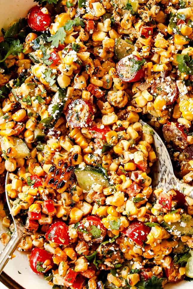 up close of two spoons serving esquites (Mexican Corn Salad ) in a white bowl garnished with cotija cheese, cilantro and lime wedges