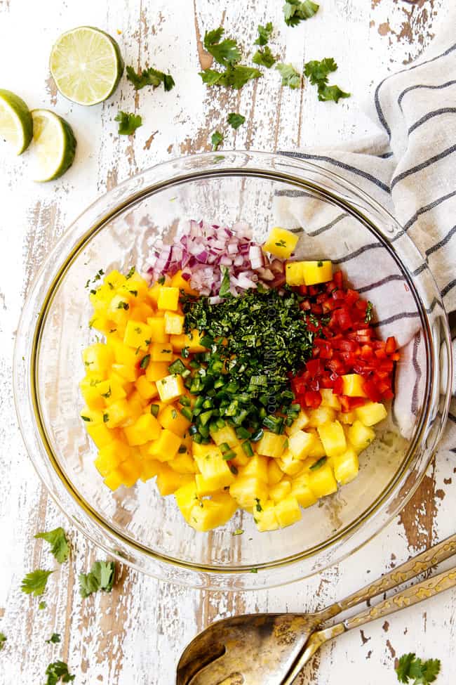 showing how to make top view of pineapple mango salsa by adding pineapple, mangos, red onions, red bell peppers, cilantro and jalapeno to a glass bowl