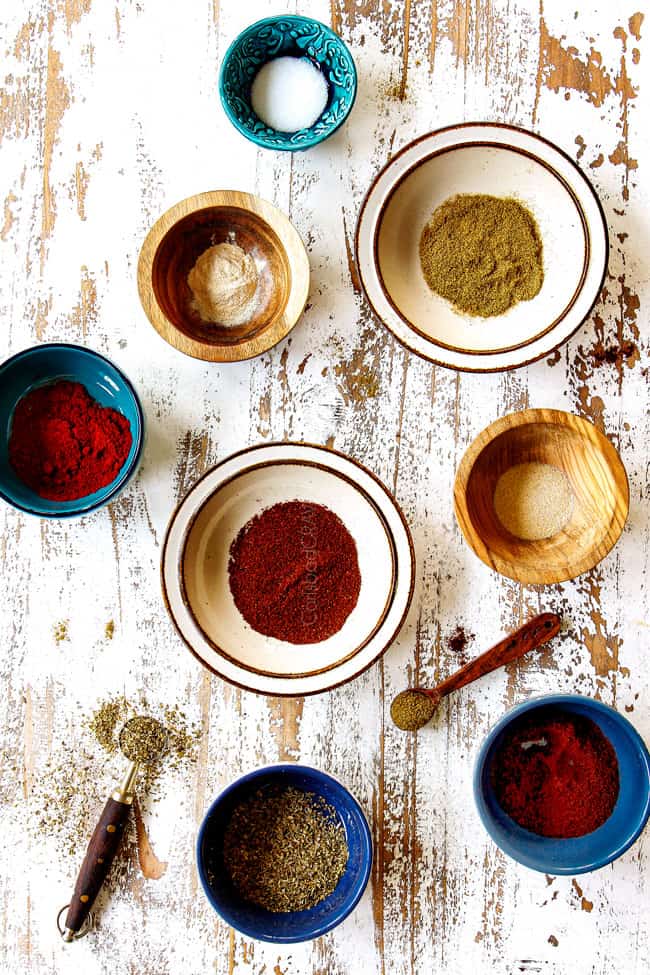 Ingredients for homemade fajita seasoning in bowls o a white wood board: chili powder, ground cumin, smoked paprika, garlic powder, onion powder, salt, pepper and cayenne pepper.