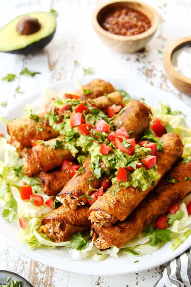 flautas recipe stacked on a white plate with shredded lettuce, guacamole and chopped tomatoes