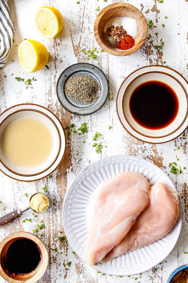 top view of showing how to make grilled chicken marinade by laying out ingredients in bowls: balsamic, lemon juice, Dijon, oregano and ketchup and chicken breasts on a white plate