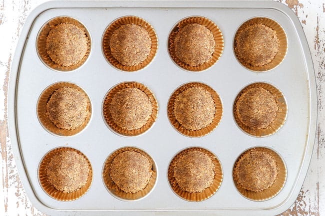 process shot showing how to make mini pumpkin cheesecakes by filling 12 muffin tins with graham cracker crumbs to make a crust