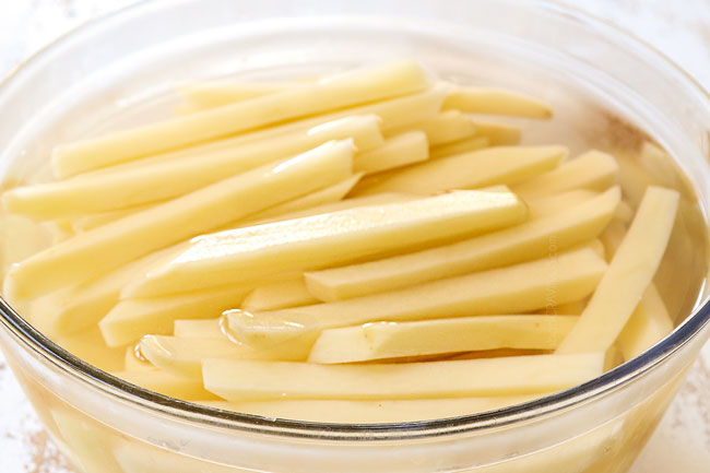 showing how to make poutine by soaking cut potatoes in cold water in a glass bowl