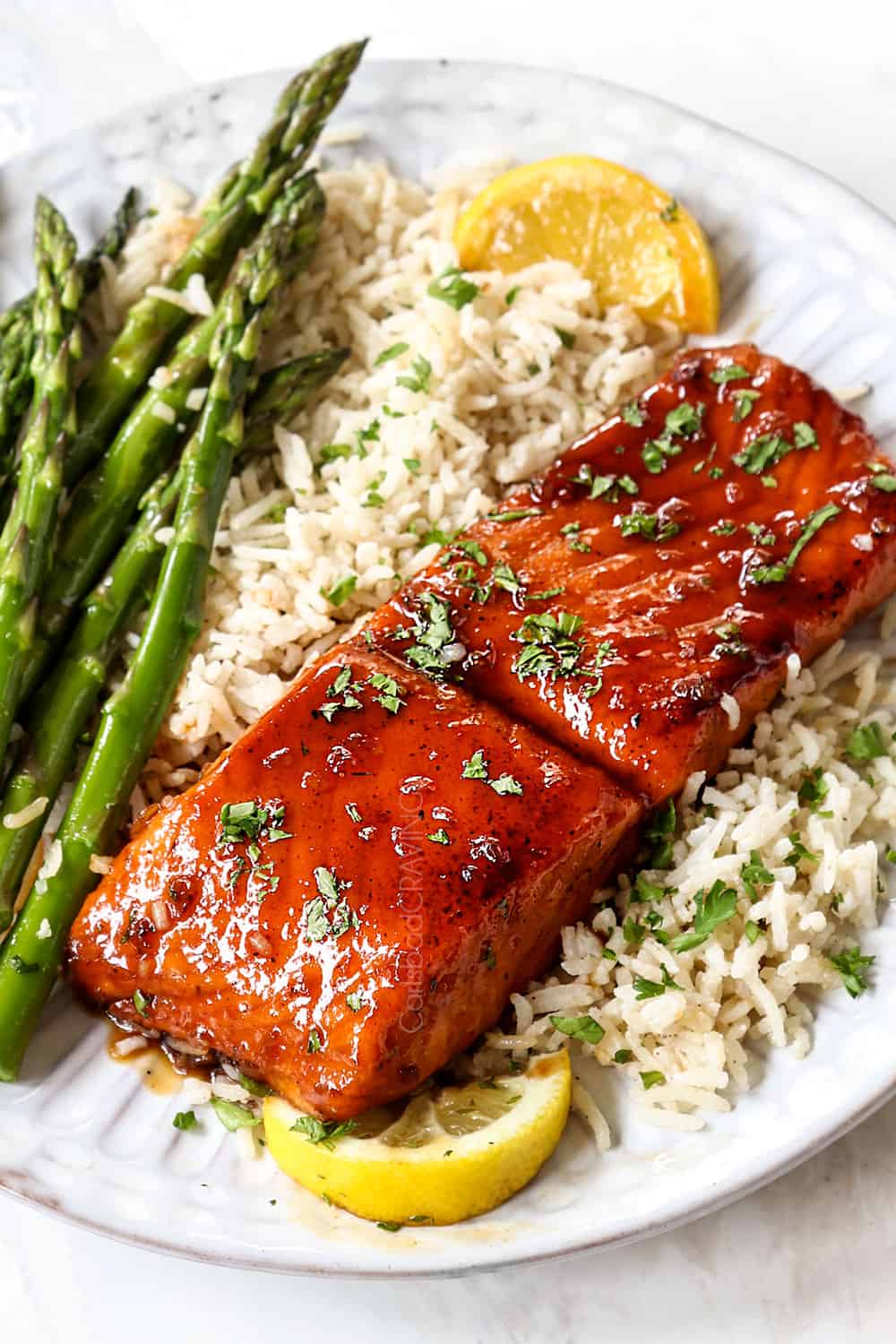 showing how to serve honey garlic glazed salmon by serving on a white plate with jasmine rice, asparagus and lemon wedges garnished by chopped parsley,