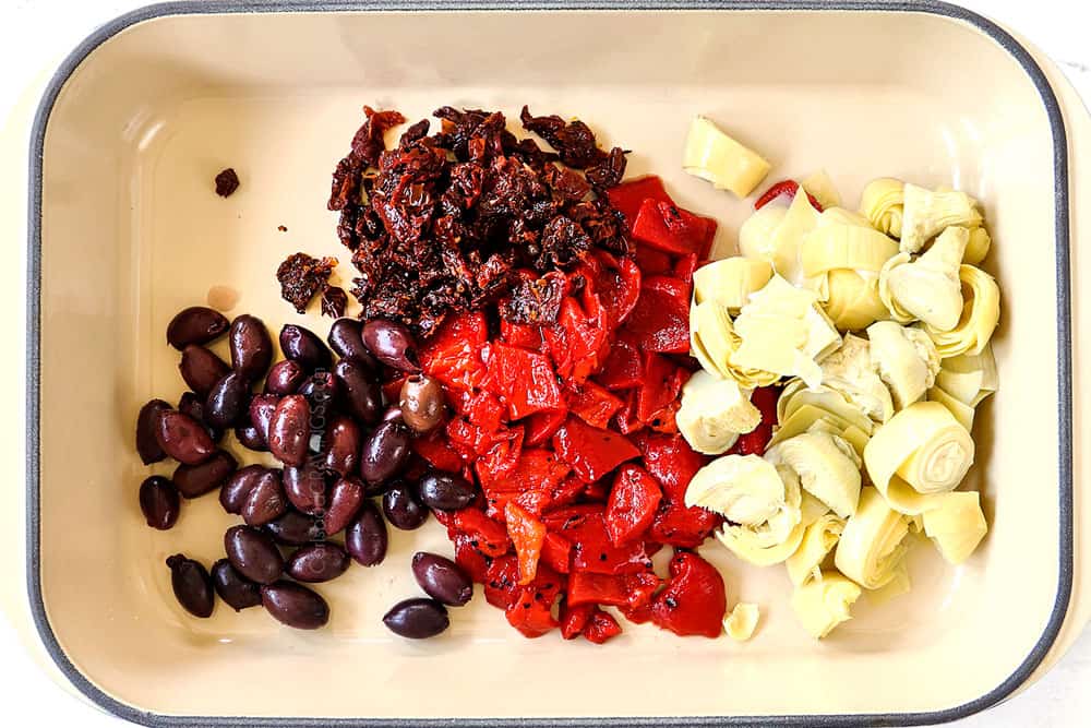 showing how to make Greek chicken by adding sun-dried tomatoes, Kalamata olives, artichokes and roasted red bell peppers to a baking dish