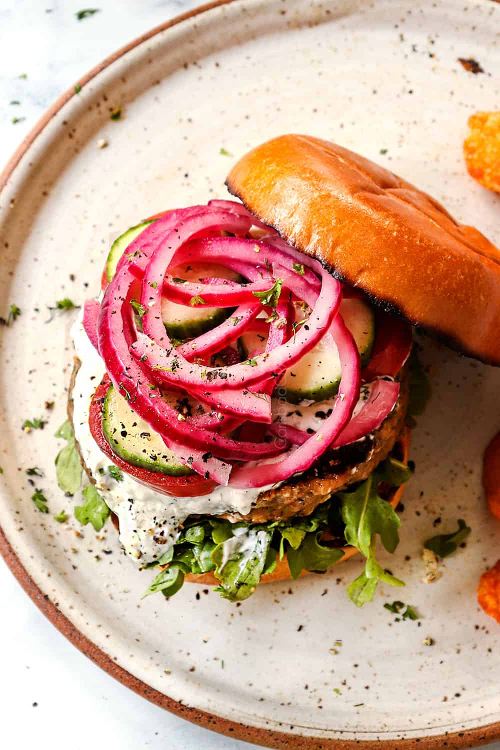 showing how to make Greek burger recipe by layering buns with arugula, lamb burger patties, tzatziki, tomatoes, cucumbers and red onions