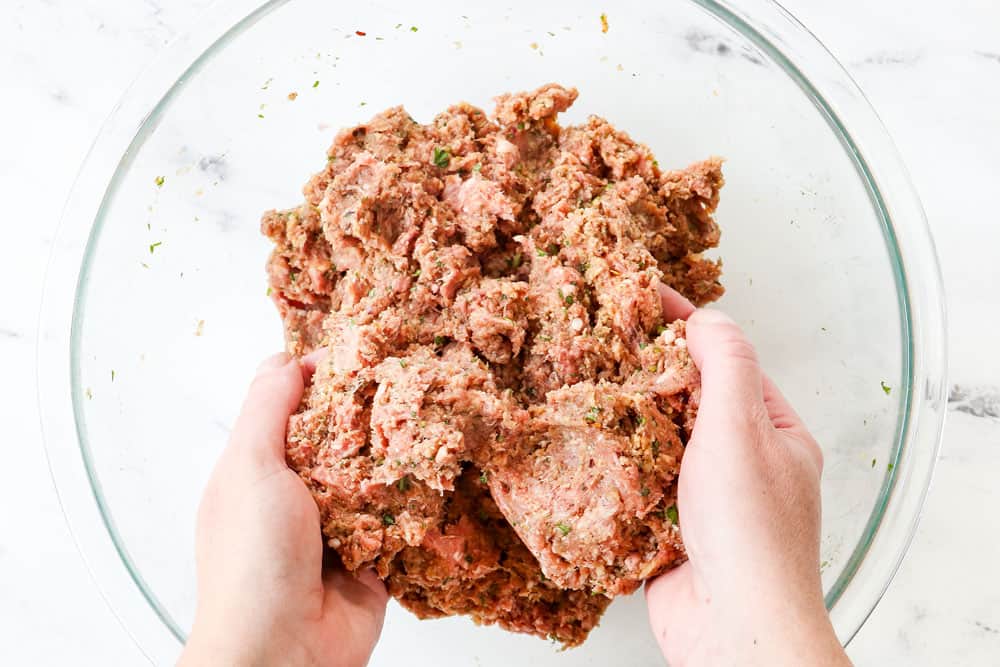 showing how to make Greek Meatball recipe (Keftedes) by mixing ground lamb, ground beef, eggs, breadcrumbs, lemon juice, garlic, onion, basil, dill, oregano, mint to a glass bowl with two hands