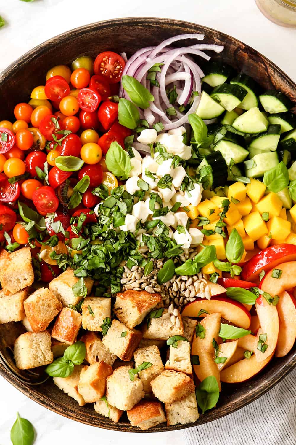 showing how to make Panzanella recipe by adding tomatoes, onions, cucumbers, bread, basil and mozzarella to a large serving bowl