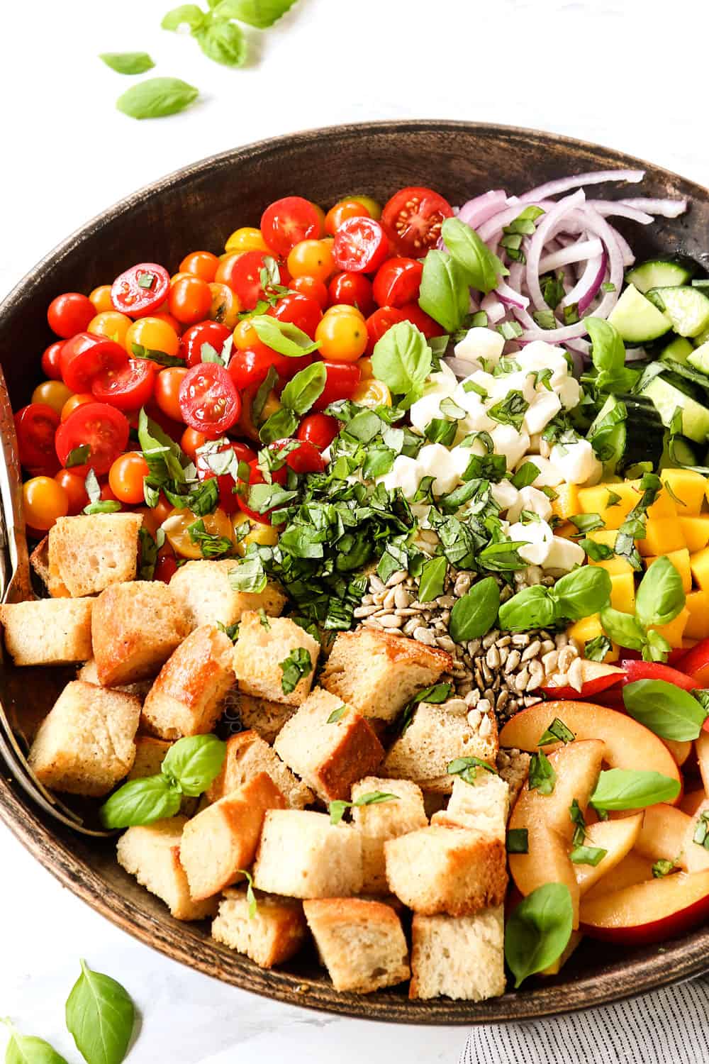 side view of showing how to make panzanella by adding the ingredients to a wood bowl: cubed bread, tomatoes, onions, cucumbers and basil