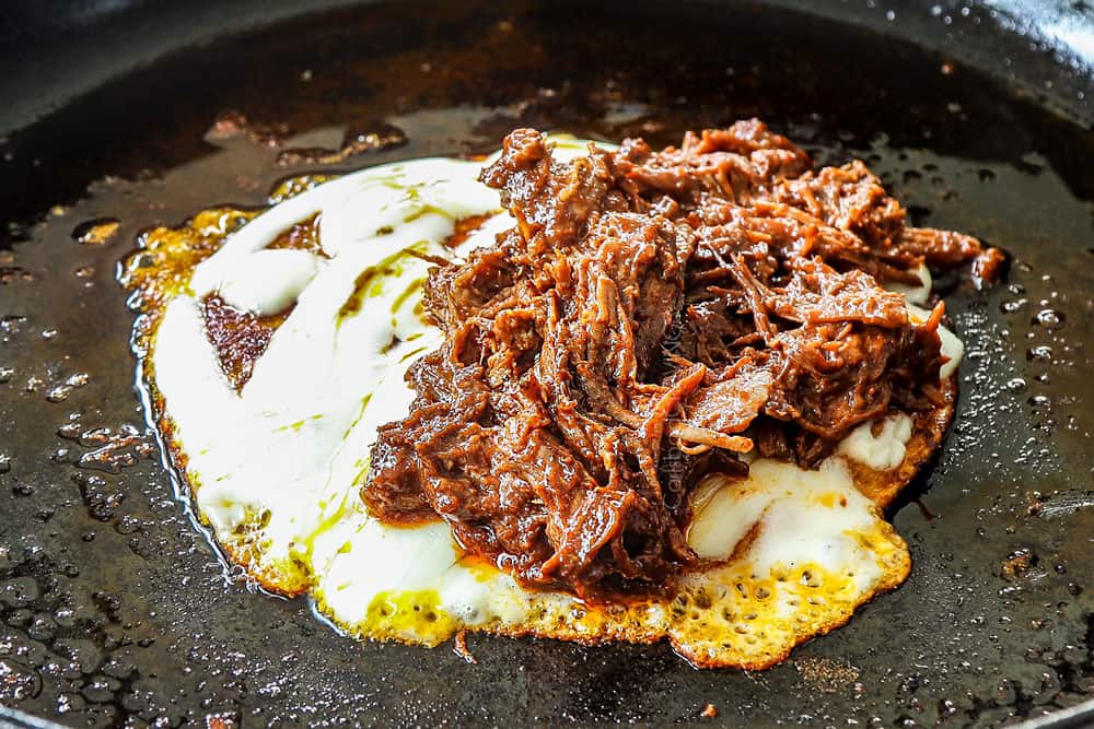 showing how to make quesa birria tacos by adding cheese to the tortilla and topping with birria de res