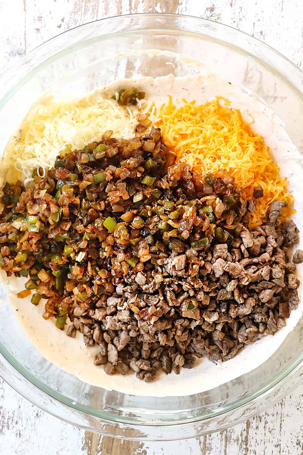 showing how to make philly cheesesteak dip by adding the cream cheese, provolone, onions and bell peppers to a glass bowl
