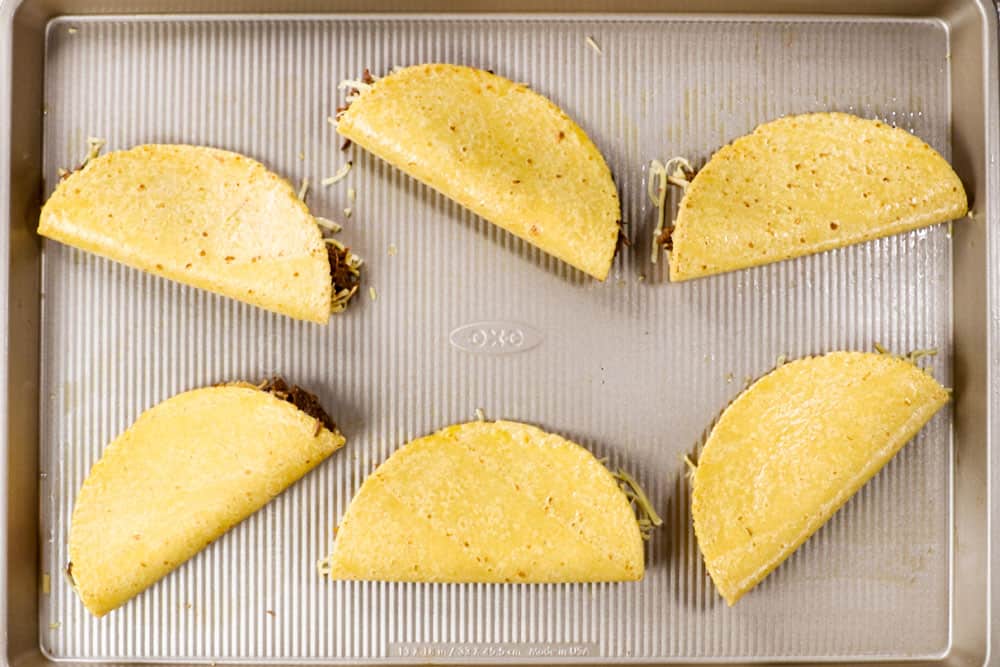 showing how to make shredded beef taco recipe by folding the corn tortillas over to form tacos on a baking sheet before baking