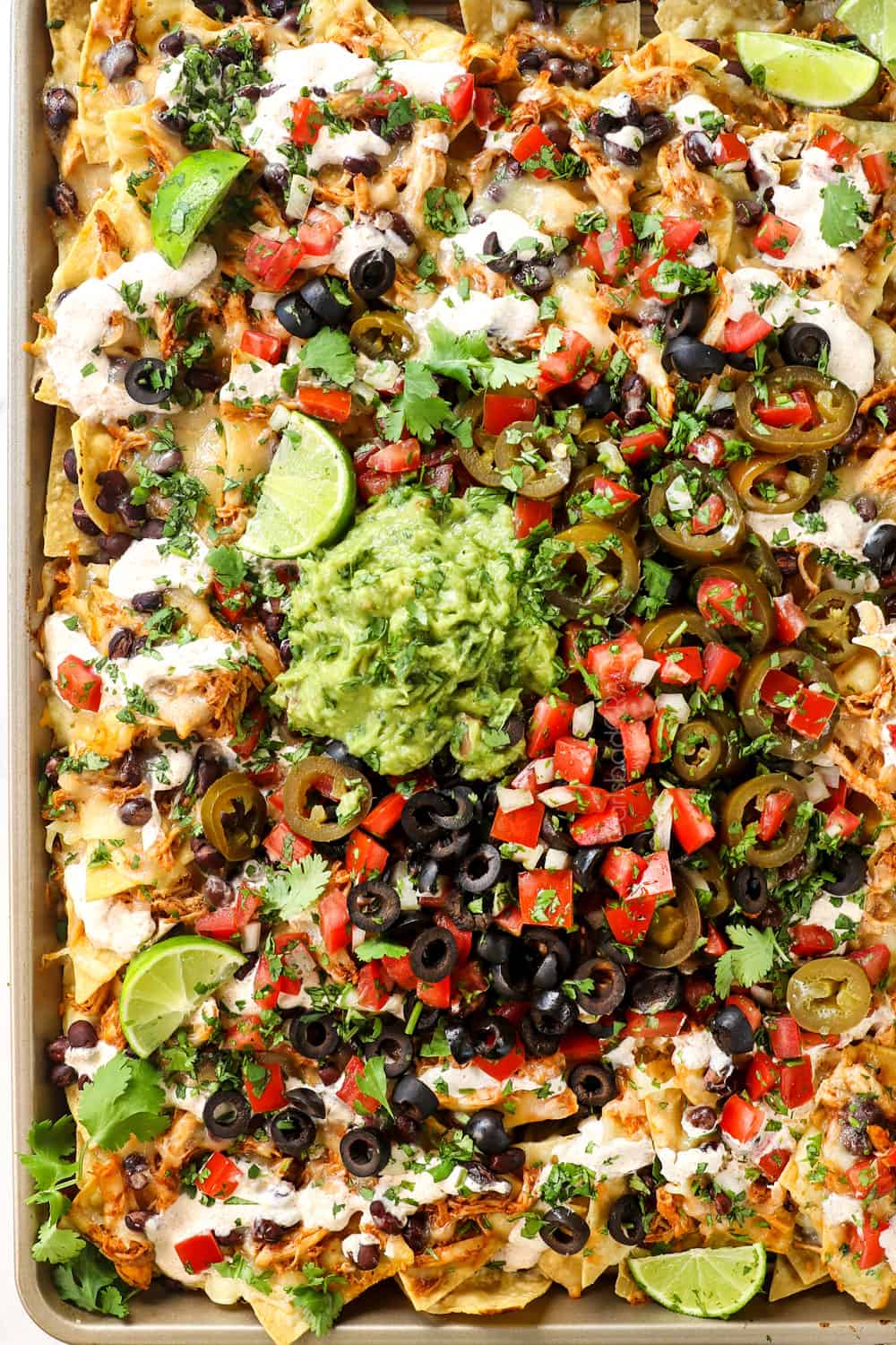 showing how to make chicken nachos by adding guacamole, olives, pico de gallo, jalapenos and sour cream on top of chips, beans and cheese
