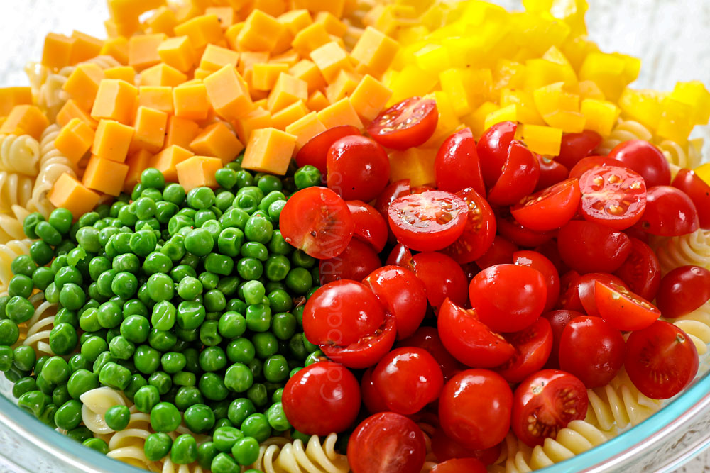 showing how to make bacon ranch pasta salad recipe by adding pasta, tomatoes, peas, bell peppers and cubed cheese to a bowl