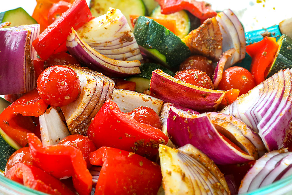 showing how to make Greek Chicken Kabobs by marinating bell peppers, onions, tomatoes and zucchini in chicken kabob marinade in a bowl